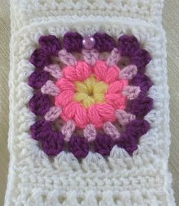 Etui pour téléphone portable fait main au crochet : en bride, tout blanc avec une fleur au coeur jaune, pétales rose, parme et violet. Fermé par une perle ronde parme.